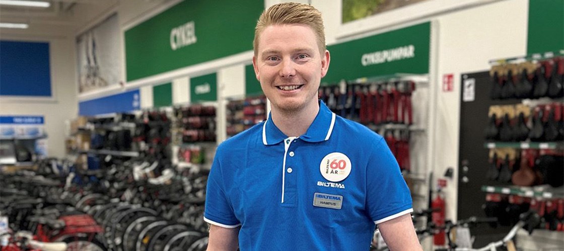 Male employee in blue Biltema uniform smiling in front of store shelves with products in the background.