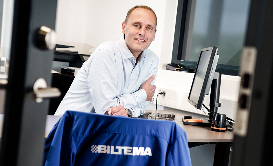 Mand i lyseblå skjorte sidder smilende ved skrivebord med computer på kontor, med Biltema-jakke hængende over stoleryg