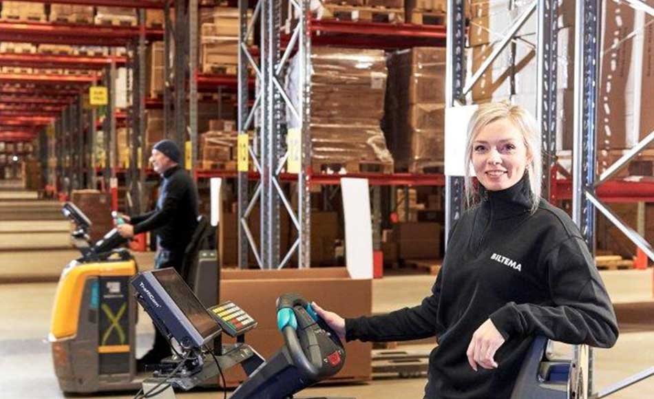 Biltema-medarbejdere arbejder i lagerhal med høje reoler og pallevarer, kørende på elektriske palleløftere.
