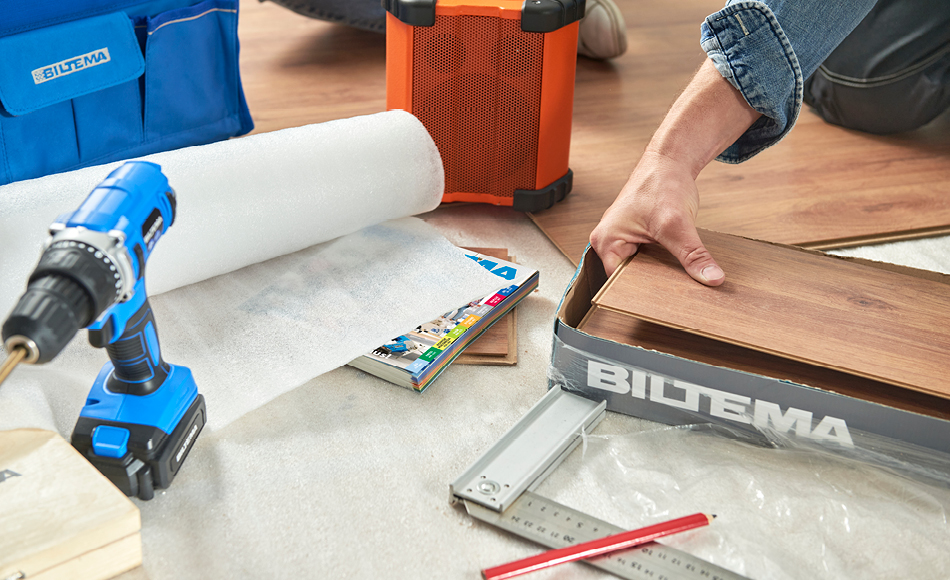 Person lægger laminatgulv med Biltema-emballage, værktøj, skruemaskine og underlagsrulle synligt på gulvet