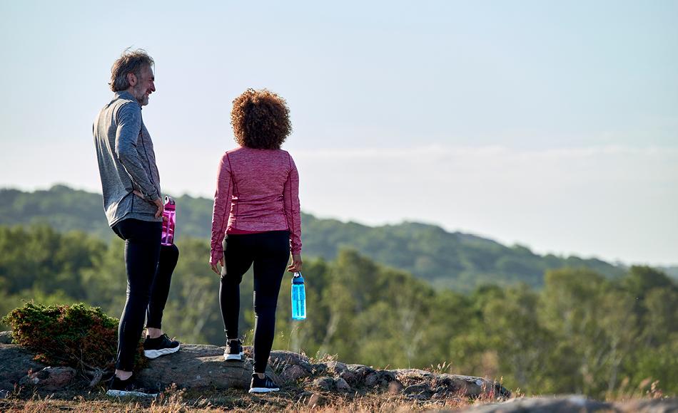 To personer i træningstøj står på en bakketop med udsigt over grønt landskab og holder farvede vandflasker