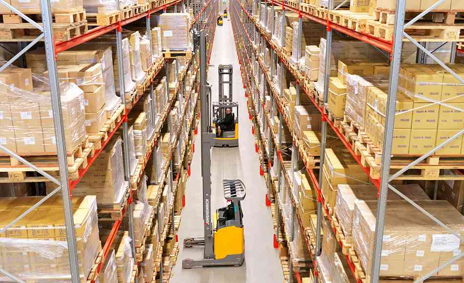 Indendørs lagerhal med høje reoler fyldt med papkasser og paller, set oppefra, hvor to gaffeltrucks manøvrerer i en smal gang mellem reolerne