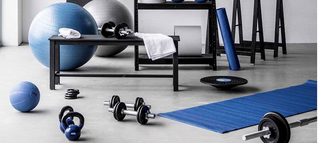 Lyst og velorganiseret hjemmegym med træningsudstyr såsom kettlebells, håndvægte, vægtstang, træningsbolde og yogamåtte. Udstyret er pænt placeret på en reol og rundt i et moderne træningsrum med hvide vægge og betongulv. En håndskrevet "Squat Challenge"-tavle bidrager til den motiverende træningsatmosfære.