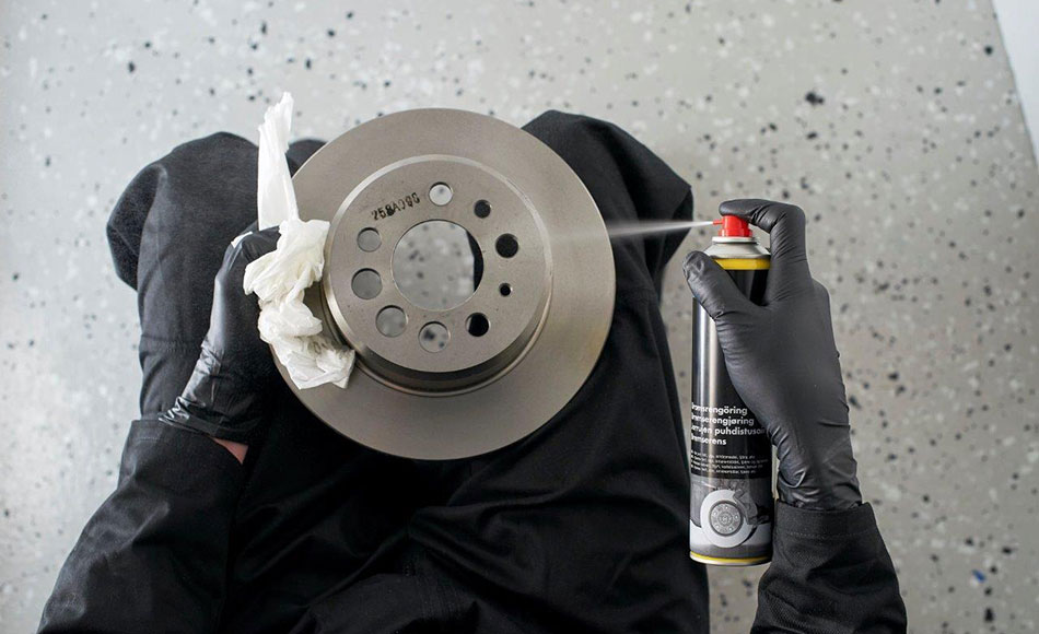 Person med sorte handsker rengør bremseskive med sprayrens og papirklud på værkstedsgulv med grå nuancer.