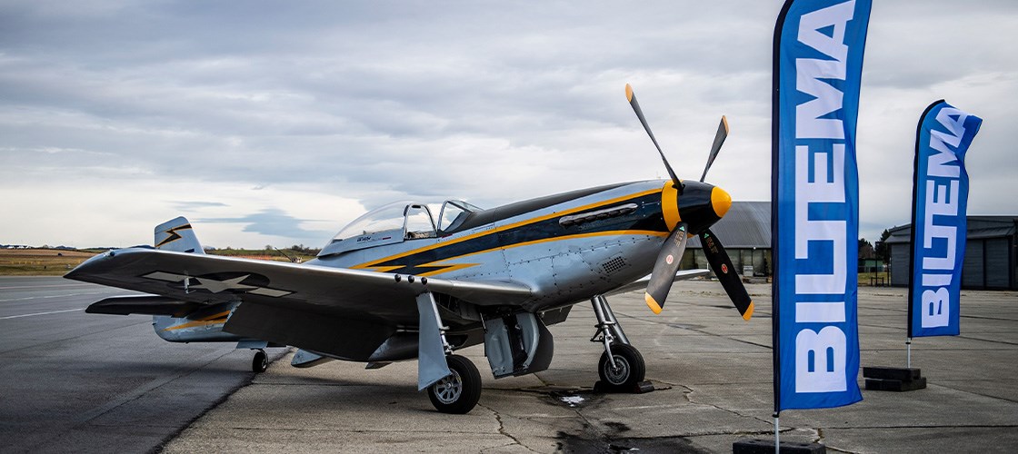 Historisk jagerfly af typen P-51 Mustang parkeret på landingsbane ved siden af Biltema-flag under overskyet himmel.