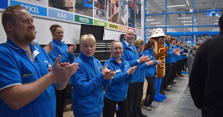 Biltema-medarbejdere i blå uniformer står på række og klapper under butiksåbning, med maskot i pølse-kostume iblandt dem