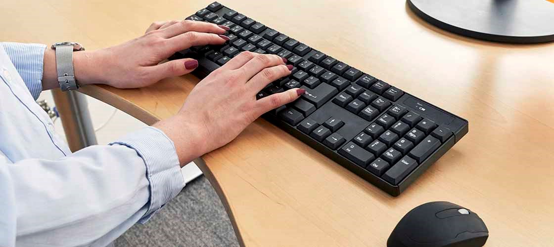 Et par kvindehænder arbejder på et sort tastatur. På hendes venstre arm bærer kvinden et sølvfarvet ur. Til højre, på bordet, kan man se dele af en computermus.