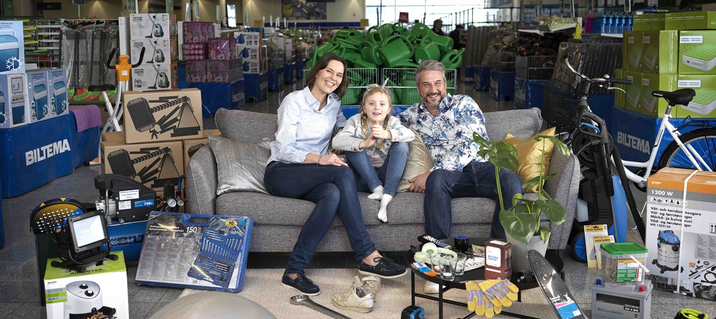 Smilende familie sidder i en sofa midt i Biltemas varehus omgivet af produkter til hus, have og værksted.