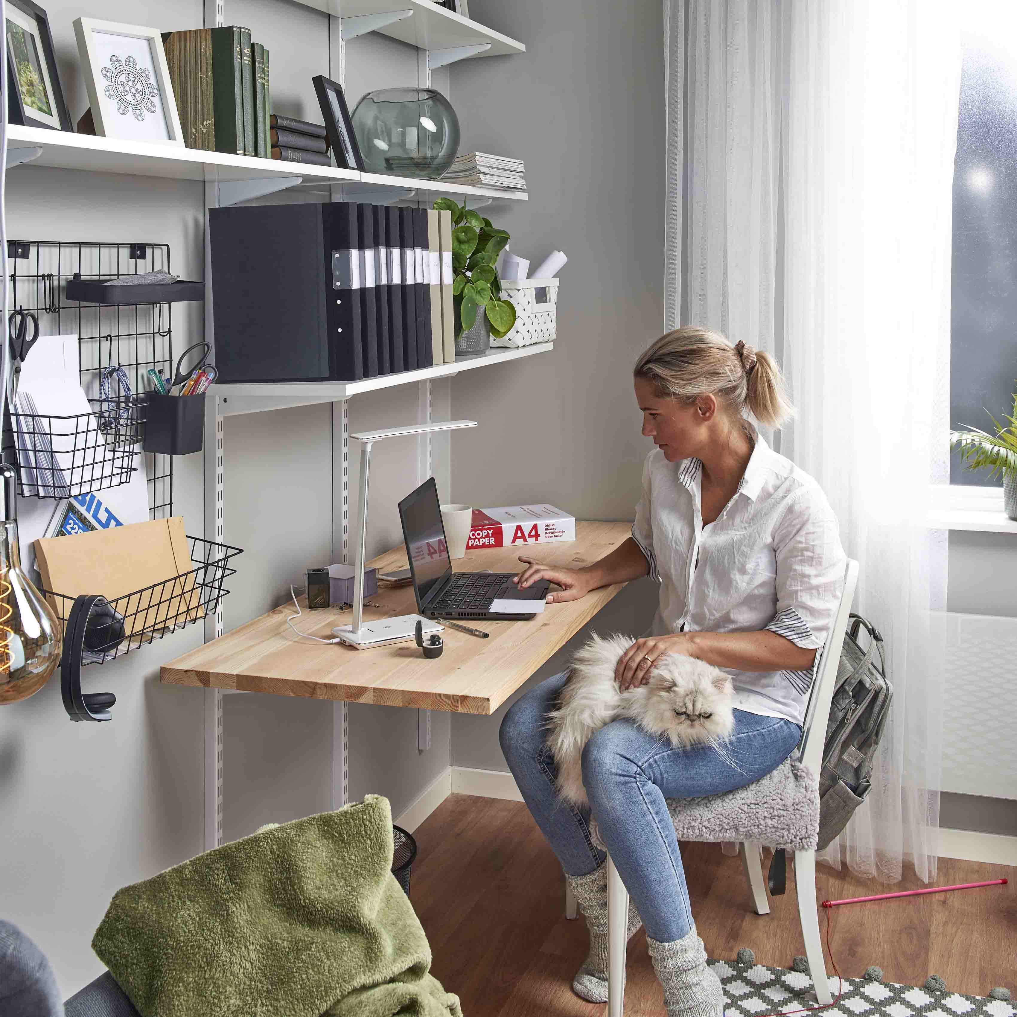Kvinde arbejder på bærbar computer i hjemmekontor med hvid langhåret kat på skødet og kontorartikler fra Biltema.