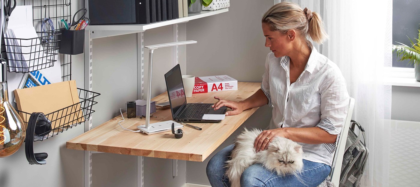 Kvinde arbejder på bærbar computer i hjemmekontor med hvid langhåret kat på skødet og kontorartikler fra Biltema.