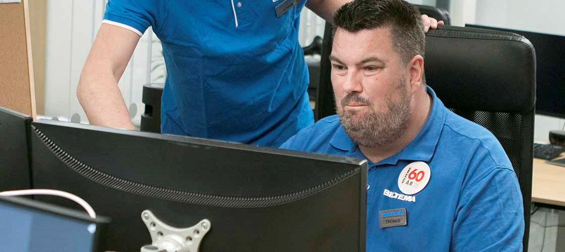 An employee in blue work clothes sits in front of a computer screen and works, while a colleague stands next to him and looks at the screen with him.