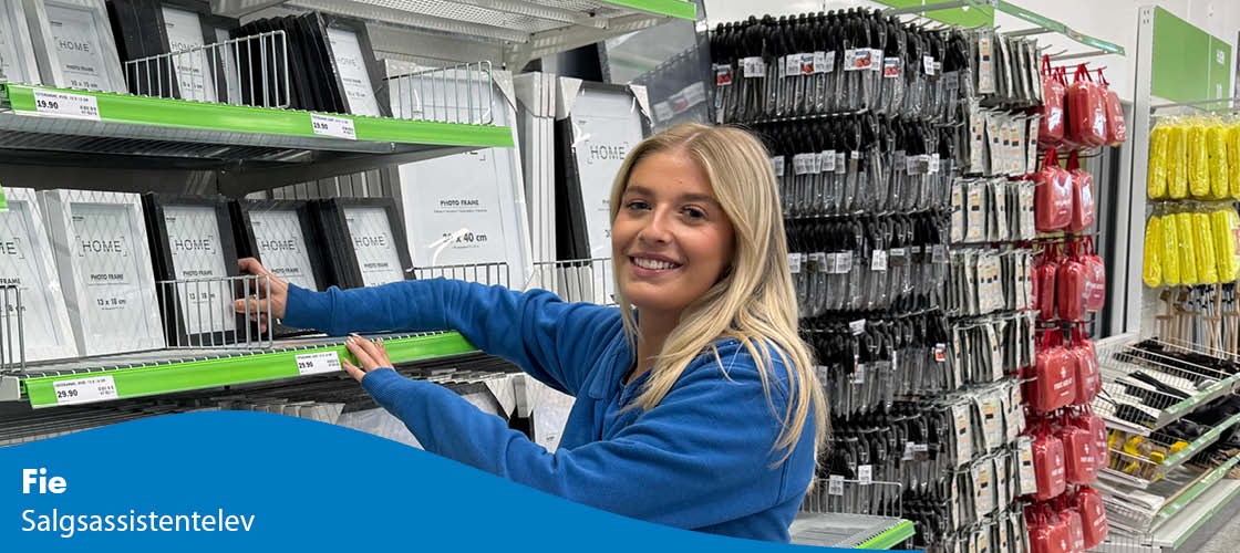 Medarbejder i Biltema-butik ved hylder med billedrammer og varer.