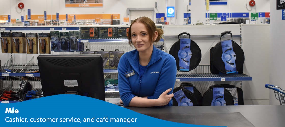Employee at the checkout in a Biltema store with goods on shelves in the background.