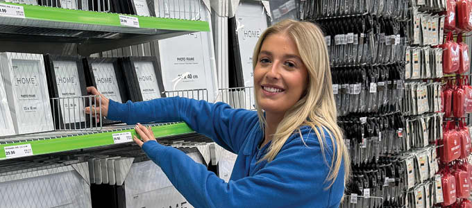 Medarbejder i Biltema-butik ved hylder med billedrammer og varer.