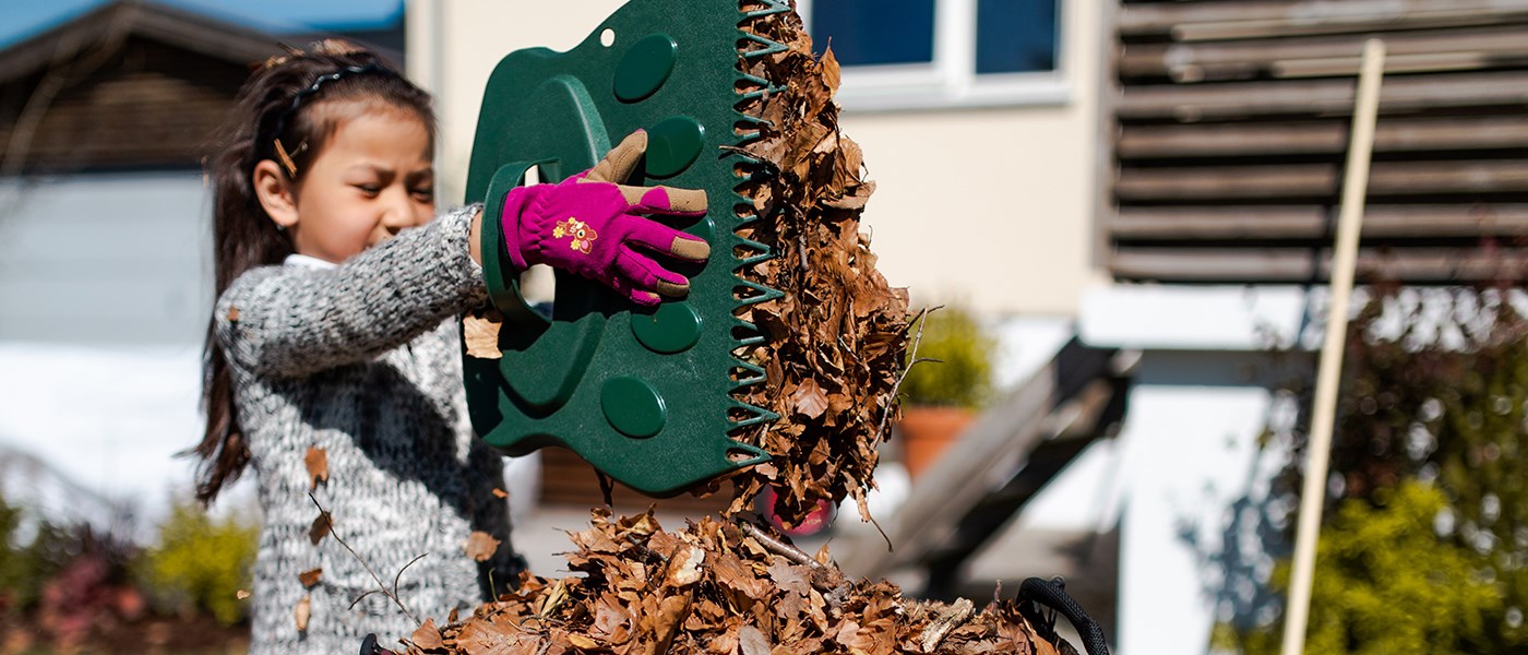 Pige iført grå striktrøje og pink handsker samler efterårsblade op med et grønt plastikhaveværktøj foran et hus i solskin.