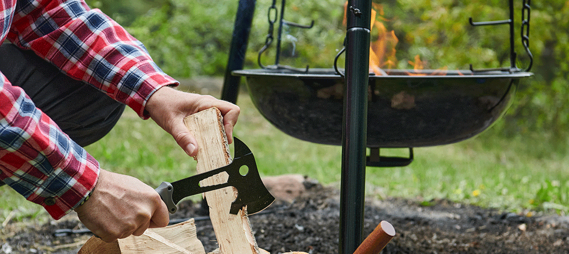 Person in a checked shirt chopping firewood with a small axe in front of a fire pit in the garden with a fire in it, surrounded by green nature.