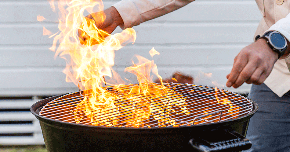 Person står ved en kulgrill med åbne flammer, hvor grillmaden tilberedes over den varme ild.
