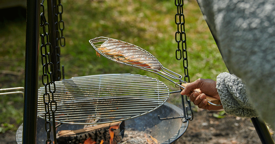 Person holder en grillrist med en fisk over et bålsted i haven, klar til at blive grillet over åben ild.