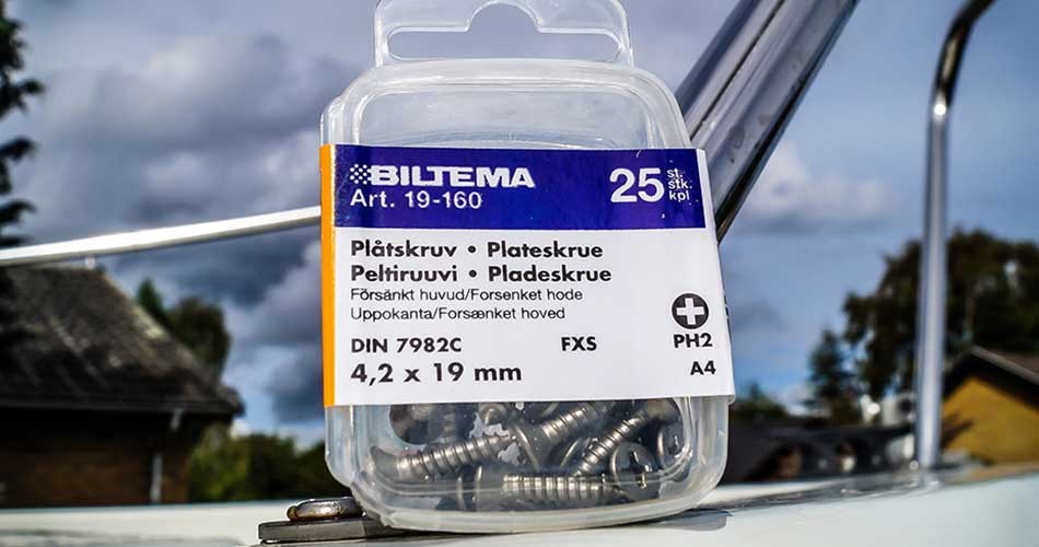 Plastbøtte med 200 stk. franske skruer i rustfri A4-stål, 6 x 60 mm, placeret udendørs med byggeri og himmel i baggrunden.