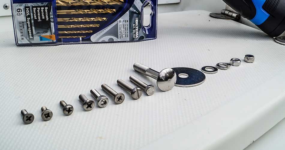 Række af forskellige skruer og bits opstillet på hvid overflade, med produktpakning og værktøj delvist synligt i baggrunden.. 