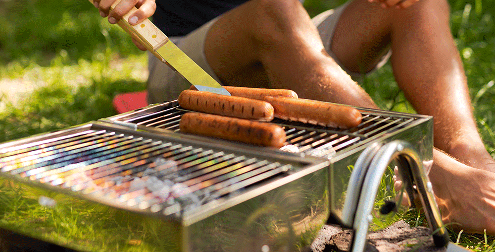 Fire pølser grilles over gløder på en kompakt camping grill i det fri, med solskin og grønt græs i baggrunden.
