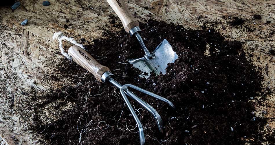 Garden tools with wooden handles, a hand cultivator and a small shovel, lying in loose, dark potting soil on a wooden base. Tools for pre-germination from Biltema.