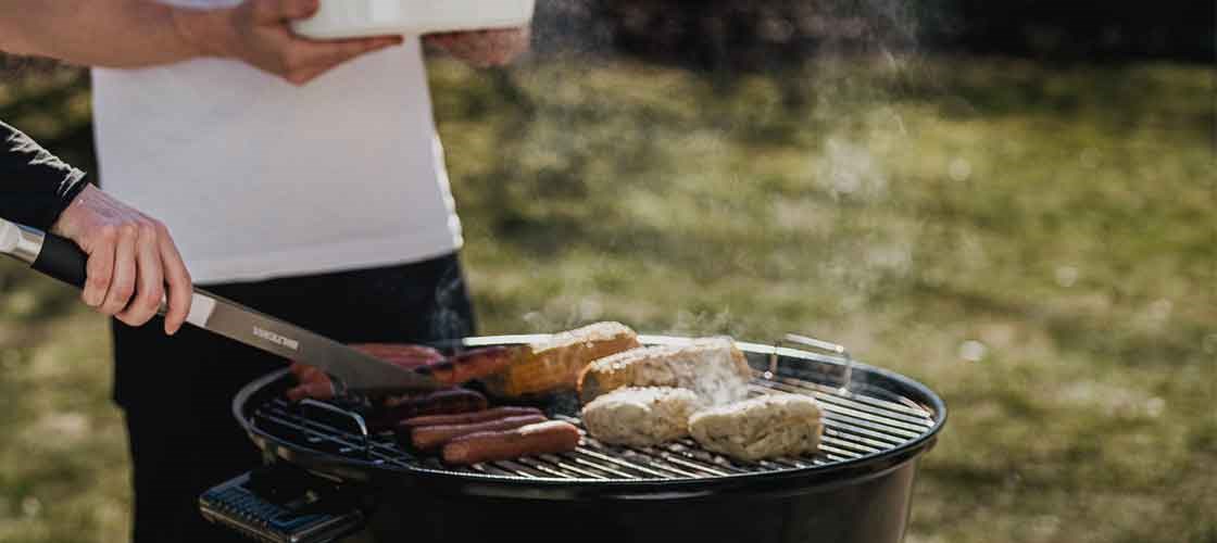 Mad tilberedes på en rund kull grill (Kuglegrill) udendørs, mens to personer står ved grillen.