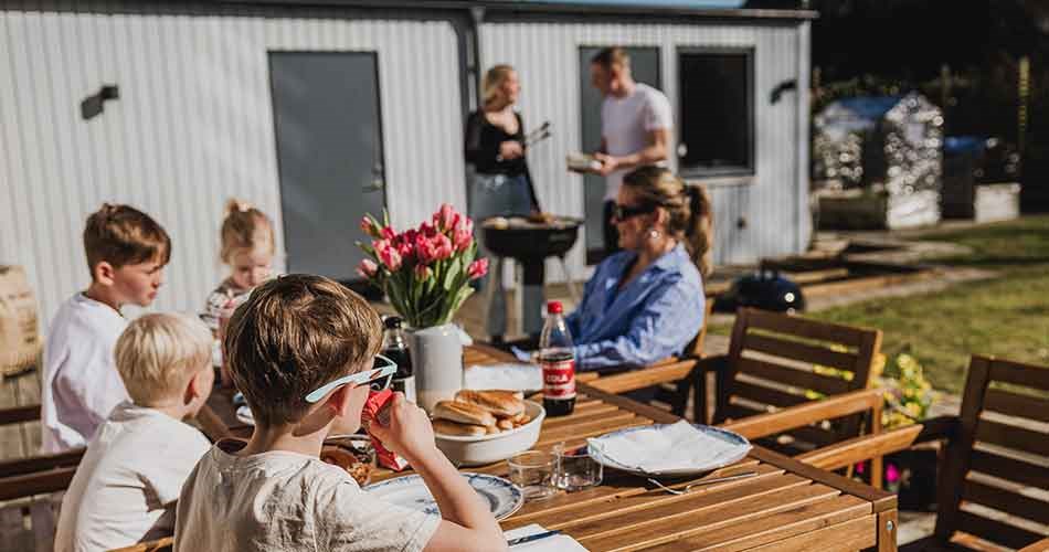 Familie samlet omkring et havebord, mens der grilles i baggrunden.