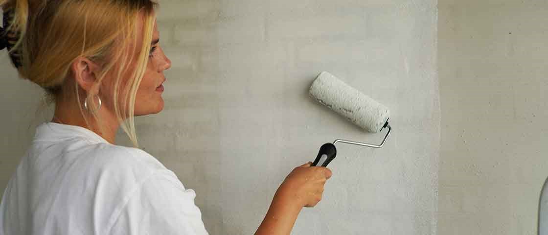 Kvindelig håndværker maler indendørs væg med Biltema malerrulle og hvid vægmaling under boligrenovering.