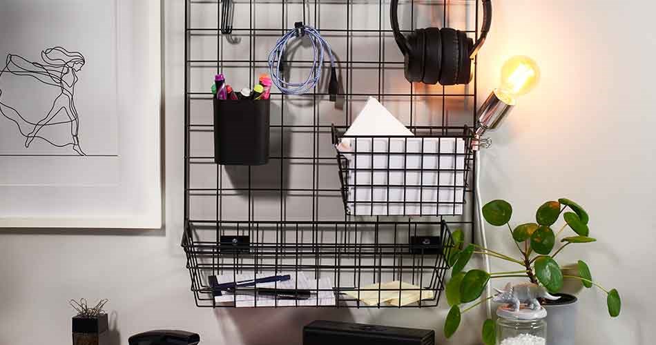Vægophængt metalgitter med opbevaringskurve, høretelefoner, kabler og lampe ved hjemmekontor.
