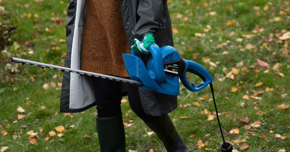 Person wearing gardening gloves and wellies uses hedge trimmer to prune autumn garden.