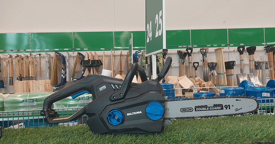 Electric chainsaw displayed in front of a row of gardening tools in a shop, with green grass as a base and green back wall with Biltema logo in the background.
