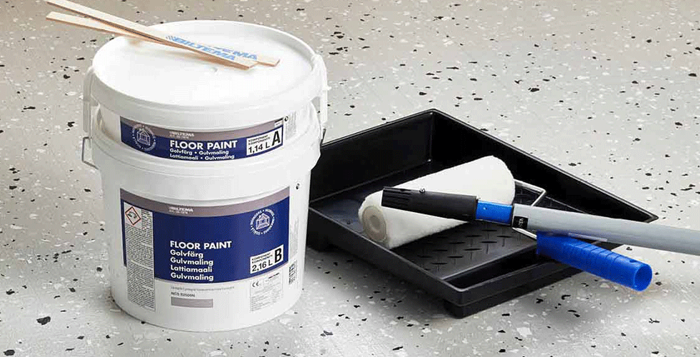 A bucket of epoxy floor paint from Biltema sits on a light grey floor with a paint tray, roller and brush ready for use next to it.