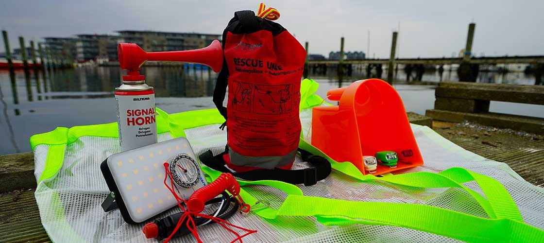 Sæt med sikkerhedsudstyr til sejlads – inkl. signalhorn, redningsline, LED-lampe med kompas, fløjte, redningsskovl og refleksvest, placeret ved kajkant med vand og både i baggrunden.