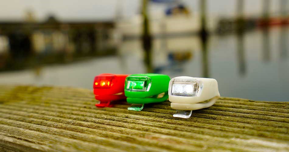 Tre små LED-lygter/nødlanterner til båd i rød, grøn og hvid placeret på en træbro med vandoverflade og havneområde i baggrunden.
