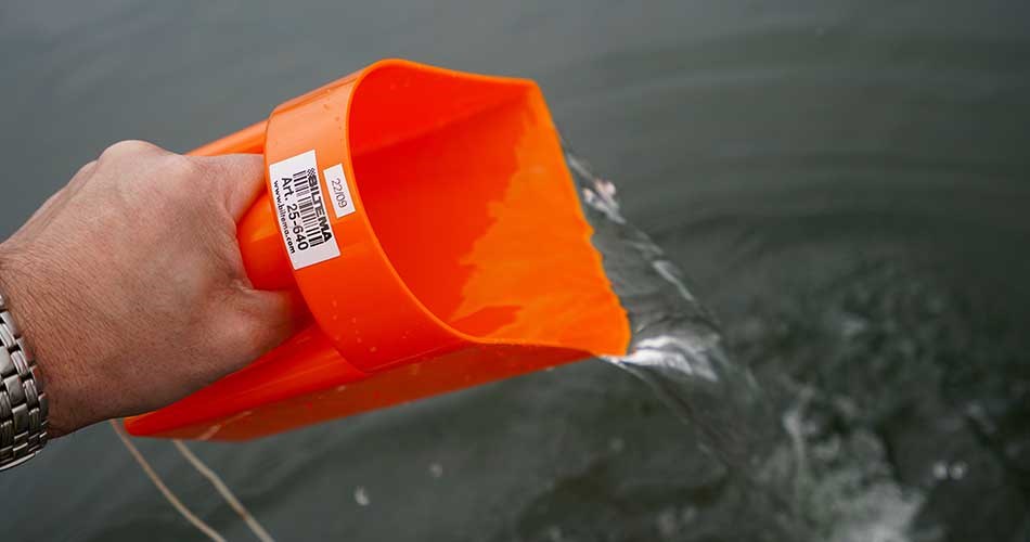 Hånd holder orange øsekar/pøs og øser vand ud af båd ved vandoverflade.