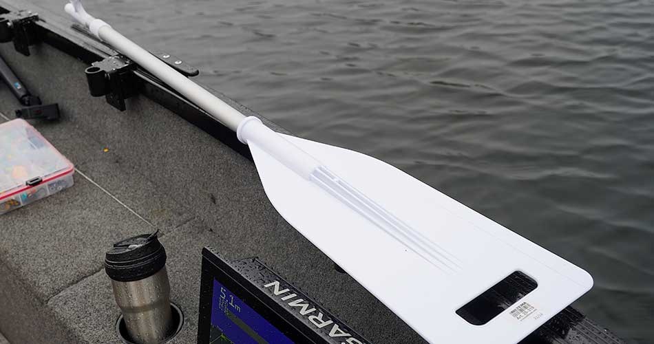 Hvid kombineret bådshage og åre fra Biltema liggende på kanten af en båd med gråt skrog, med stille vand i baggrunden.