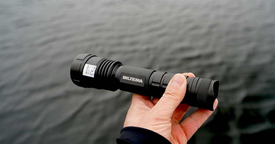 Hånd, der holder en sort lommelygte med Biltema-logo over en mørk vandoverflade. Lygten kan bruges til at signalere til andre både til havs.