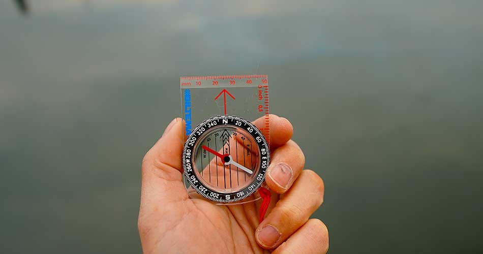 Hånd, der holder et gennemsigtigt kompas med rød og sort nål foran en rolig vandoverflade.