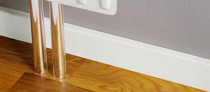 Hvid fodliste monteret langs væg under radiator med rør gennem udskæring.