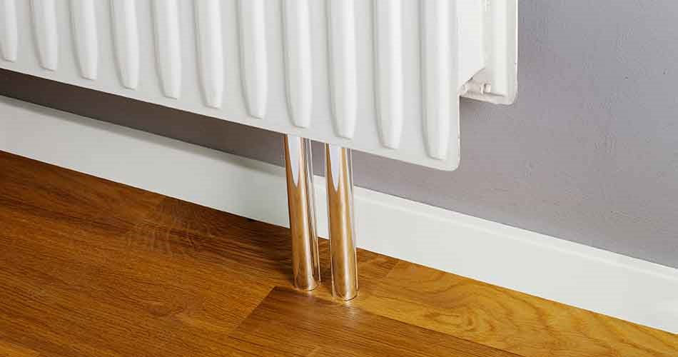 Hvid fodliste monteret langs væg under radiator med rør gennem udskæring.