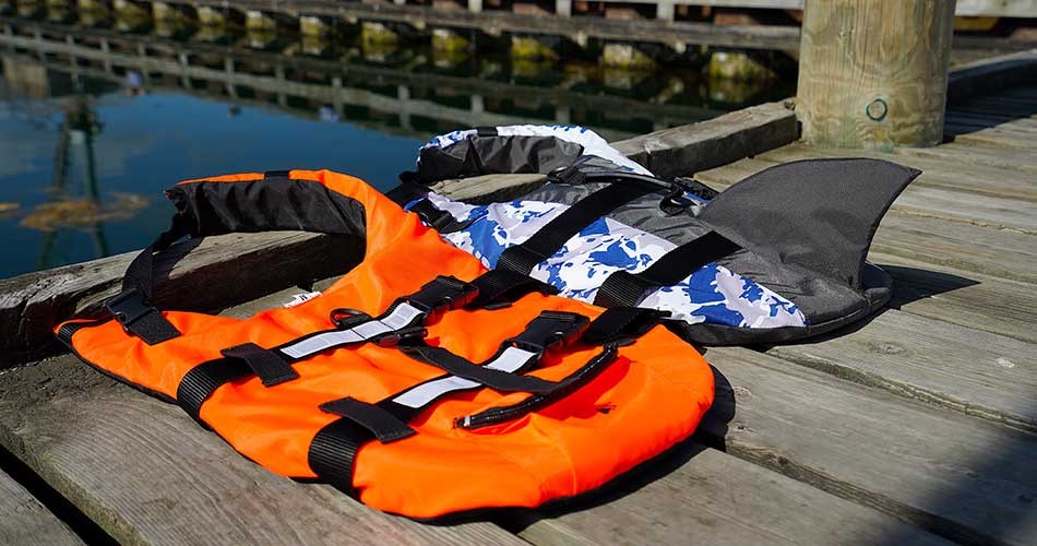 Two dog life jackets lying on a jetty - one in orange with black buckles and one in blue and white camouflage with shark fin.