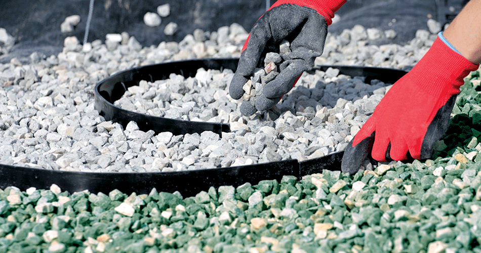 Red-gloved hands lay small decorative stones around a black border in a garden, with green and white stones distributed in different sections.