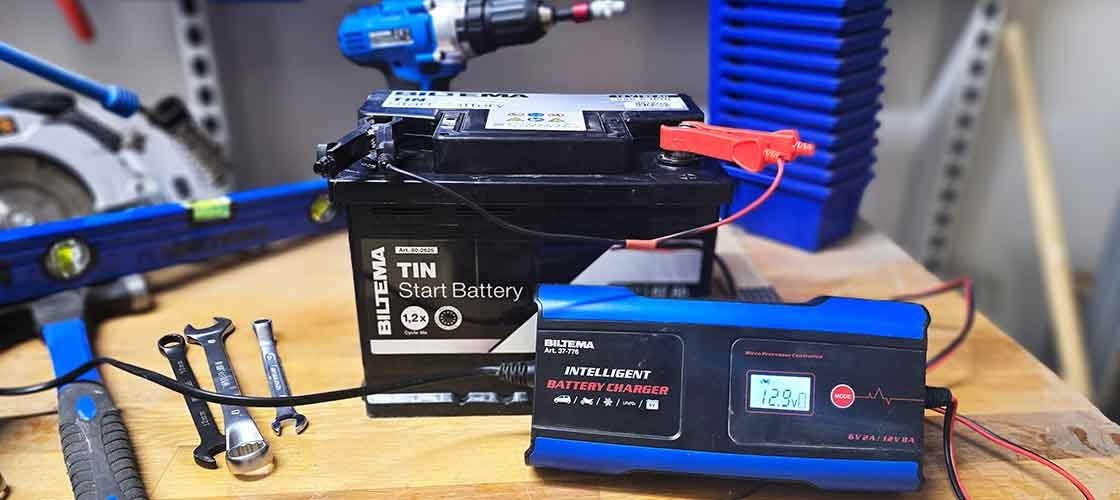 Car battery connected to a charger on a workbench with tools beside it.