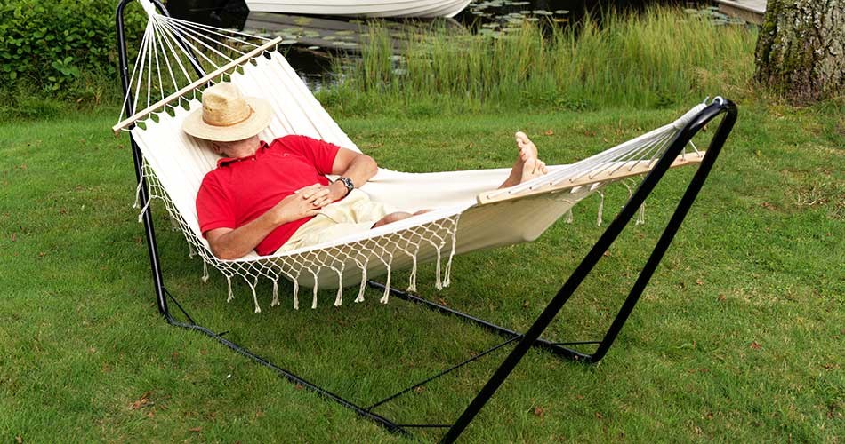 Person in a red T-shirt and straw hat relaxing in a hammock with a metal frame from Biltema on a green lawn.