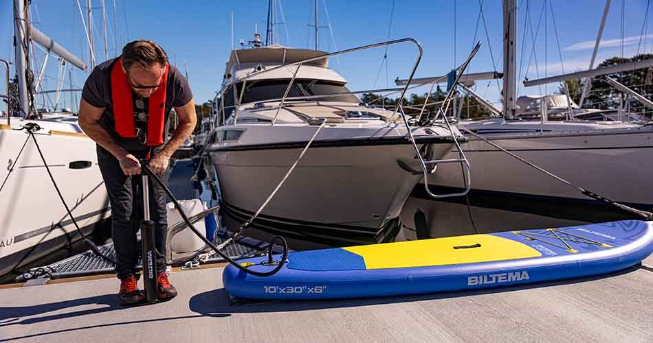 En person iført redningsvest pumper et oppusteligt SUP-board op på en marina, klar til en dag på vandet.