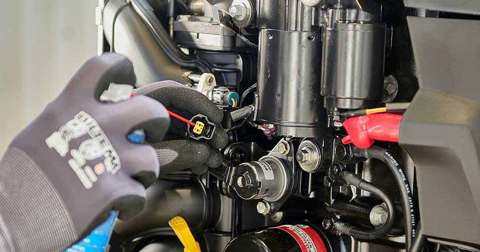 Close-up of a person wearing grey work gloves performing maintenance on a black boat engine with visible wires and components.