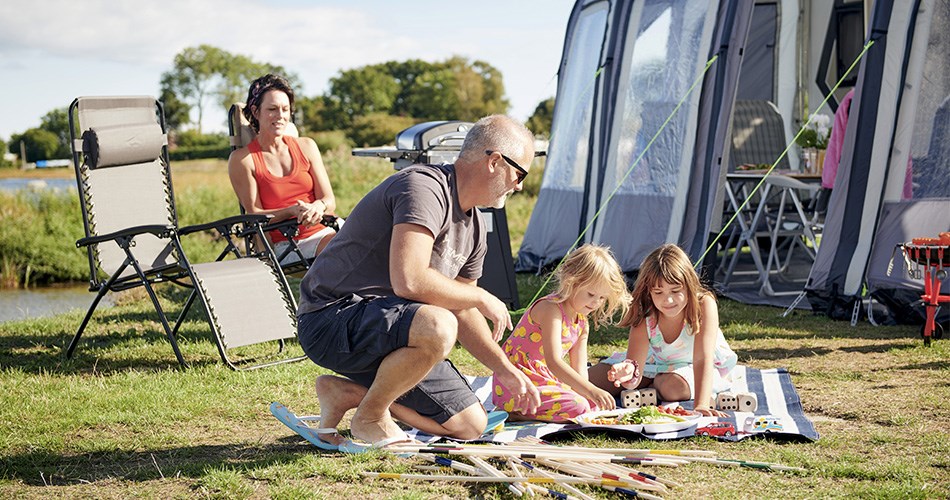 Family enjoying camping life at a campsite - two children playing on a blanket, a man sitting by them and a woman relaxing in a camping chair in the background.