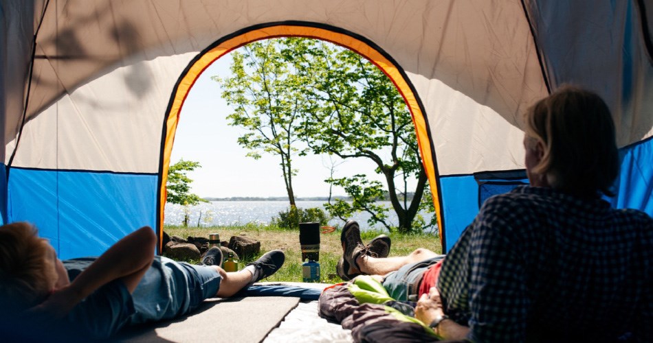 View of lake through opening in tent - two people relaxing with their feet stretched out, surrounded by nature and trees.