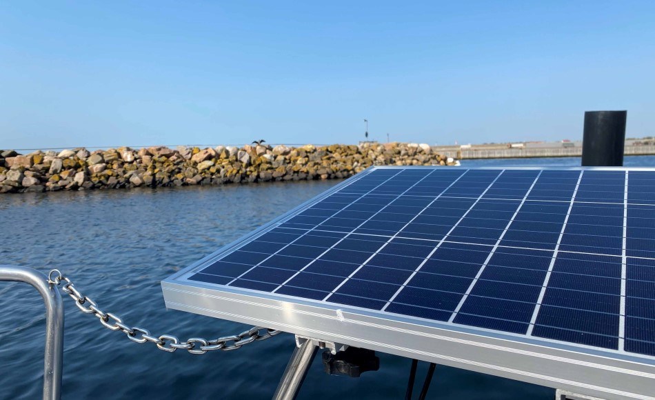 A solar panel is mounted on the rail of a boat lying still in a harbour. In the background is a breakwater made of large stones and a clear blue sky, indicating sunny conditions - ideal for solar energy.